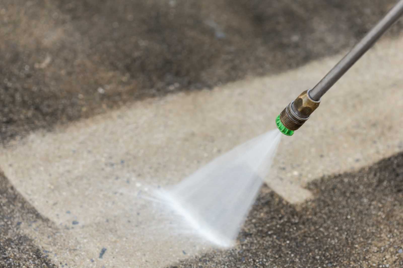 An image of a pressure washer spraying water at a concrete surface to clean it. The water stream is powerful and directed towards the ground, creating a splash and removing dirt and debris from the surface